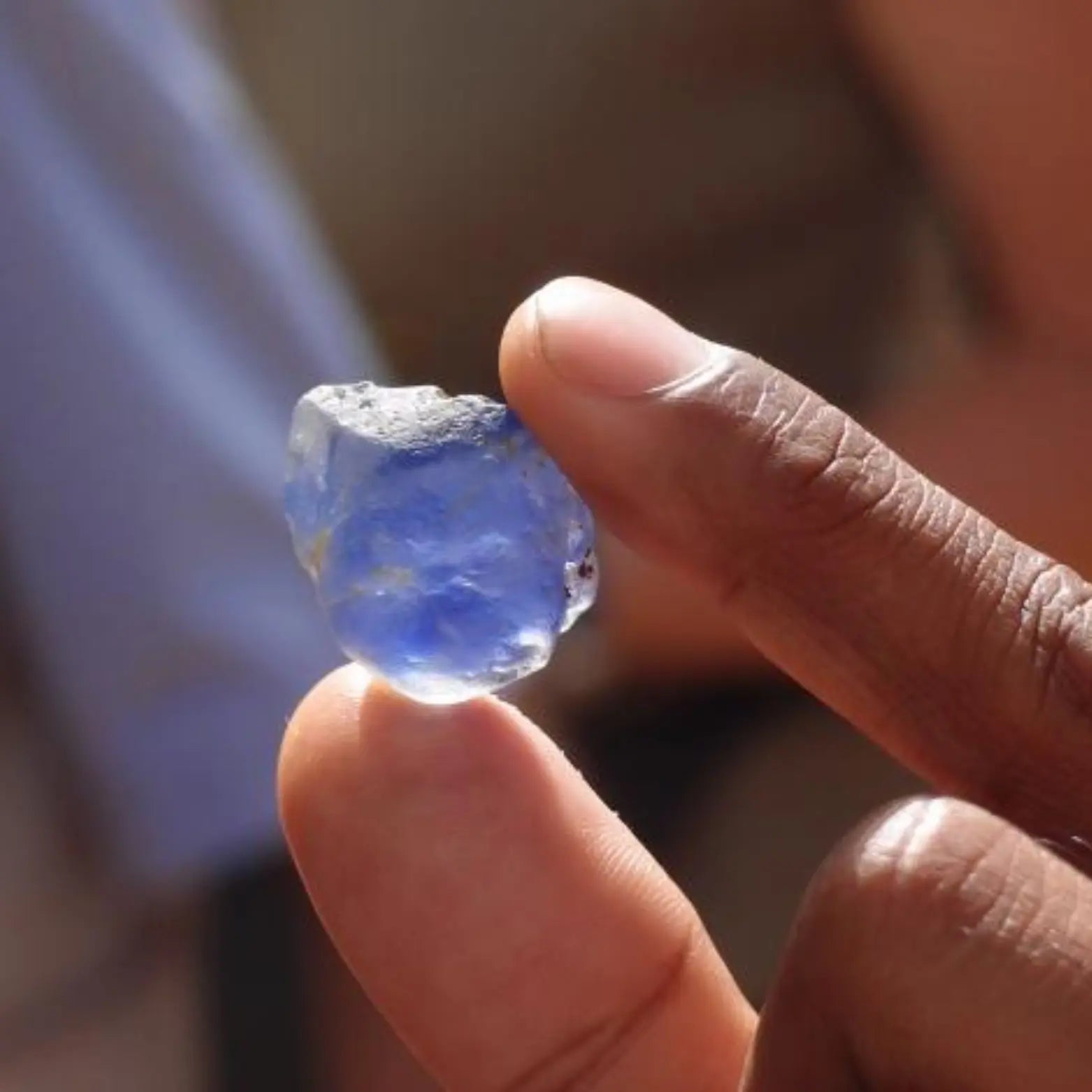 Gemme brute d’un bleu intense tenue entre les doigts d’un artisan lors de son extraction. Cette pierre précieuse naturelle, encore non taillée, illustre le processus d’extraction éthique et le travail minutieux des artisans avant la transformation en gemme de joaillerie. - mael gems