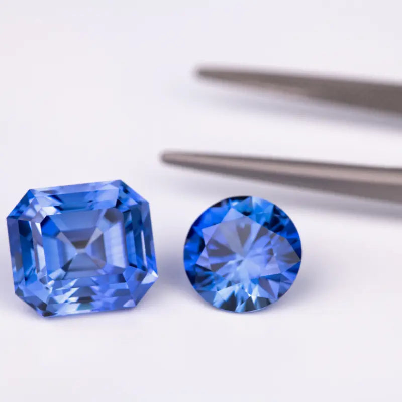 Deux saphirs taillés, l’un en taille émeraude et l’autre en rond brillant, posés sur un fond blanc avec une pince à gemmes en arrière-plan. Mael Gems bijoux sur mesure.