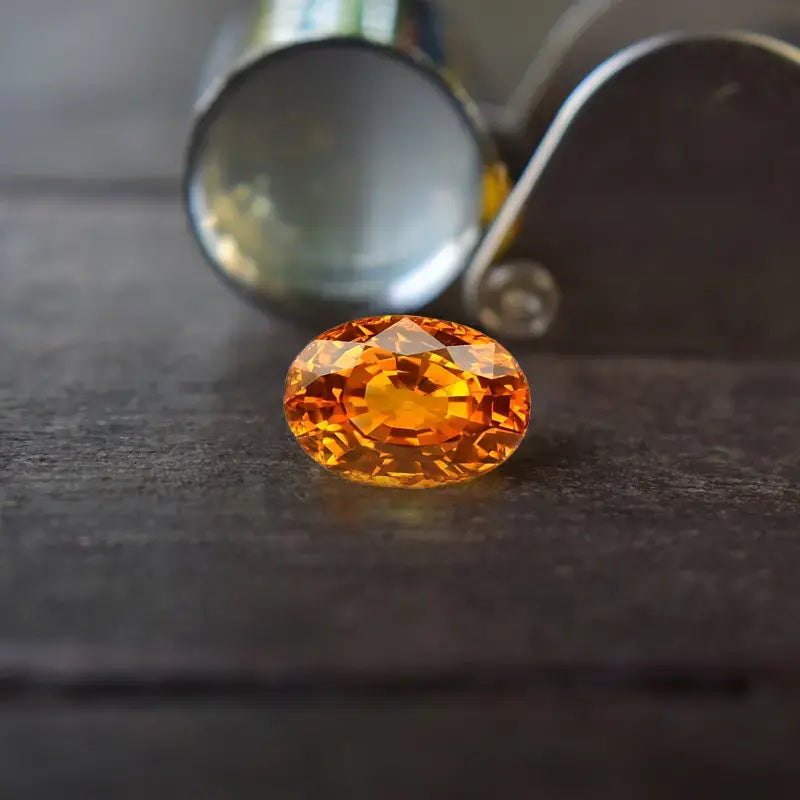 Superbe saphir orange taillé en ovale reposant sur une surface sombre, accompagné d’une loupe de gemmologue en arrière-plan. Cette pierre précieuse éclatante est idéale pour la haute joaillerie et les collectionneurs passionnés de gemmes rares.